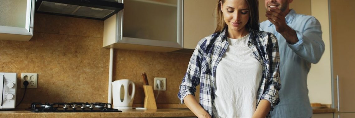 Young man covering girlfriends eyes with hands and surprising her in the kitchen at home. Loving couple cooking breakfast in the morning